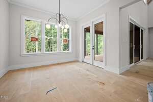 An unfurnished room with bare plywood floors, large windows with trees visible outside, a modern chandelier, and glass double doors leading to a covered porch. White trim and light-colored walls are present throughout.
