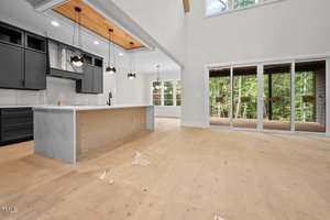 Unfurnished kitchen and living area with light wood floors, dark cabinets, a large kitchen island, pendant lights, and sliding glass doors leading to a screened porch overlooking trees. Construction debris is on the floor.