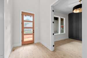 A view of a modern entryway with a wooden front door featuring glass panels, adjacent to a room with a large window, dark accent wall, and a ceiling light fixture, both with light-colored walls and floors.