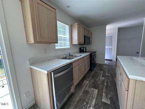 A modern kitchen with light wood cabinets, white countertops, stainless steel appliances, double sink, and wood-patterned flooring. A window is above the sink and a door is visible at the end of the room.