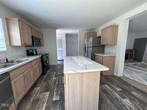 Modern kitchen with light wood cabinets, stainless steel appliances, a central island with marble countertop, and wood-look flooring. Adjacent room is visible through an open doorway, showing some flooring materials.