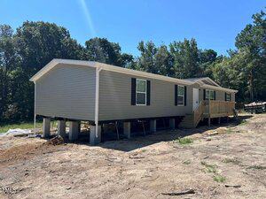 A beige manufactured home at 212 Ellington Path, Henderson, is elevated on concrete piers on a cleared lot with bare soil. It features white trim, green shutters, and a wooden porch with steps. Trees are visible in the background under a clear blue sky.