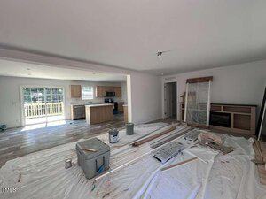 A room under renovation with plastic sheets, paint cans, and construction materials on the floor. The kitchen area with cabinets and appliances is visible in the background, and large sliding glass doors let in natural light.