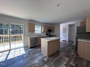 Modern kitchen with light wood cabinets, stainless steel appliances, an island in the center, and a sliding glass door leading to a deck. The room has gray-tiled flooring and white walls.