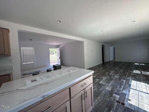 View from a kitchen counter looking into an open-plan living area with gray tile flooring, white walls, and natural light coming from windows and an open front door. Tools and papers are on the counter and floor.