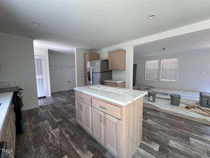 A kitchen with light wood cabinets, a center island, and stainless steel appliances. Nearby, an adjoining room has plastic coverings and construction materials, indicating ongoing renovations. The floor has a wood-look finish.