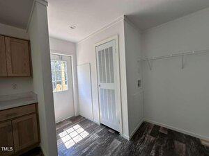 A laundry area with a closet door, a wire shelf, hookups for a washer and dryer, wood-look flooring, and an exterior door letting in sunlight next to kitchen cabinets on the left.