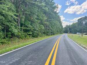 A two-lane asphalt road with double yellow lines curves gently through a wooded area with tall green trees on the left and a grassy area on the right under a partly cloudy sky.