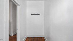 A small, empty white hallway with a wooden floor, white walls, and a black wall-mounted coat rack with several hooks. An open doorway is visible on the left side.
