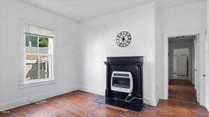 A room with white walls, a window with sunlight, wood floors, a black fireplace with a wall-mounted heater, a black clock above the mantel, and a hallway with doorways visible in the background.