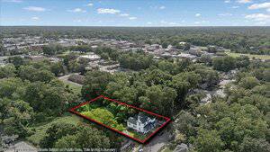 Aerial view of a residential neighborhood with a large white house and green yard outlined in red, surrounded by trees. The outline distinguishes the property from the surrounding area.