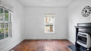A room with white walls, a wooden floor, a window with blinds, a black fireplace with a wall clock above it, and a white gas heater. The room appears unfurnished and has natural light from the windows.