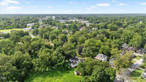 Aerial view of a suburban residential area with many trees and houses scattered among greenery; a water tower and additional buildings are visible in the background under a blue sky.