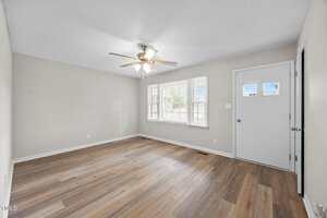 A room with light-colored walls, wood-look flooring, a ceiling fan with lights, a window, and a white front door with two square windows, viewed in daylight. The room appears empty and clean.