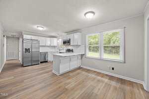 A modern kitchen with white cabinets, stainless steel appliances, a tile backsplash, and light wood flooring; adjacent to a dining area with large windows overlooking green trees outside.