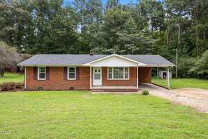 A single-story brick house at 1050 Rock Spring Church Road in Henderson features a dark shingled roof, white trim, and a covered carport on the right side. Set on a grassy lawn, it's surrounded by tall trees in the background.