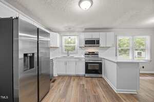 Modern kitchen with stainless steel refrigerator, stove, and microwave, white cabinets, subway tile backsplash, large windows, and light wood flooring. A ceiling light fixture is centered on the textured ceiling.
