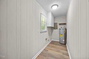 A narrow laundry room with light-colored wood panel walls, a window, a ceiling light, upper cabinets, hookups for washer and dryer, and a water heater on the right side near the back wall.