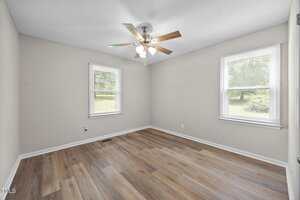 An empty room with light gray walls, two windows with blinds, a ceiling fan with lights, and wood-look vinyl flooring. Natural light enters through the windows, revealing a view of green grass and trees outside.