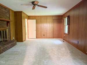 A living room with wood-paneled walls, a brick fireplace on the left, beige carpet, a ceiling fan, a window with blinds on the right, and two closed doors at the back wall. The room is empty.