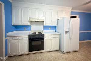 A kitchen with white cabinets, a white refrigerator, and a black electric stove and oven below a white range hood. The walls are painted blue with a white chair rail, and the floor is covered with beige tiles.