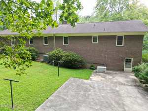 A brick house with a shingled roof, viewed from the backyard, featuring a concrete patio, a lawn with clothesline poles, air conditioning units, and a backup generator. Trees and bushes surround the yard.