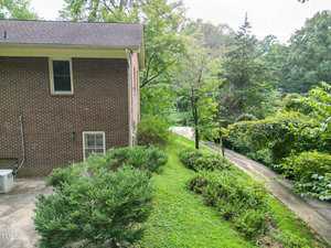 A brick house with two visible windows is shown on the left. There is an air conditioning unit, green bushes, and a grassy area leading to a driveway that curves through dense trees and vegetation.