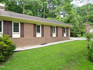 A single-story brick house with brown shutters and cream trim, surrounded by green grass and bushes. A concrete walkway runs alongside the house, and trees are visible in the background.
