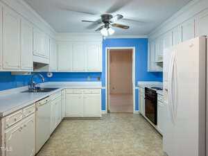 A kitchen with white cabinets, white countertops, white appliances, and a black stove. The walls are painted blue, there is a ceiling fan with lights, and the floor has a beige tile pattern. An open doorway leads to another room.