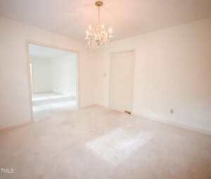 A bright, empty room with off-white walls and carpet, featuring a chandelier, an open doorway leading to another room, and a closed sliding door on one wall. Sunlight streams in through a window out of view.