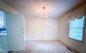Empty room with cream-colored carpet, white walls, and a chandelier hanging from the ceiling. There is a large window with drapes on the right and an open door leading to a kitchen on the left.