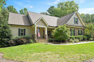 Single-story house with beige siding and stone accents, a gabled roof, and an American flag near the front porch. The yard is landscaped with green grass, shrubs, and small trees under a partly cloudy sky.