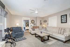 A living room with beige walls and carpet, a light-colored sectional sofa, a recliner, a walker, a wooden coffee table, framed photos on the wall, and a window with checkered curtains. A doorway leads to another room.