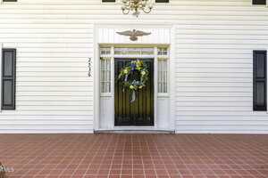 A white front door with glass panels, flanked by sidelights, features a large decorative wreath. The house has white siding, black shutters, and a red tile porch floor. House number 2536 is mounted vertically by the door.