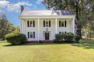 A two-story white house with black shutters on 2536 Enon Road, Oxford, features a covered front porch supported by four columns, brick steps, and green bushes beside a grassy yard and trees under a blue sky.