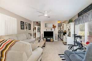 A living room with beige carpet and walls, a sectional sofa with a striped blanket, a coffee table, a fireplace, a ceiling fan, and a view into a kitchen; a walker is near a glass door to a deck.