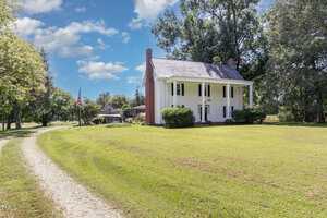 A white two-story house with large columns stands on a grassy lawn bordered by trees. A gravel driveway curves past an American flag on a flagpole, with blue sky and clouds overhead.