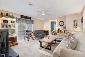 A living room with beige walls, carpeted floor, two sofas, a coffee table, TV, bookshelf, and a walker near sliding glass doors. A ceiling fan and decorative pillows are visible, and natural light enters from the left.