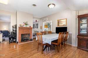 Dining area with a rectangular table and six wooden chairs, a lace tablecloth, a mirror, a china cabinet, and a brick fireplace; adjoining living room and kitchen visible in the background.