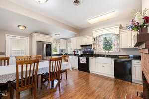 A spacious kitchen with white cabinets, black appliances, floral curtains, a wooden dining table with six chairs, hardwood floors, and a brick accent wall with a flower arrangement on the mantel.