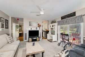 A living room with a beige sofa, a marble coffee table, and a TV on a stand. A walker sits near a sliding glass door with checkered valance. Kitchen and hallway are visible in the background.