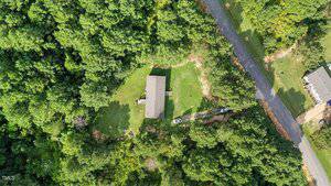 Aerial view of a single-story house surrounded by dense green trees, with a grassy yard, a driveway, and a few vehicles parked near a road on the right side of the image.