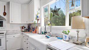 A bright kitchen with white cabinets, marble countertops, a farmhouse sink, and various kitchen utensils. A window above the sink displays greenery outside, and decorative items sit on the counter and windowsill.