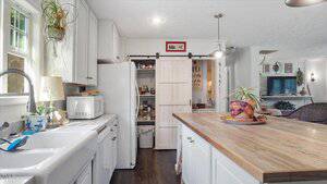A kitchen with white cabinets, a wooden island, and a farmhouse sink. A sliding barn door reveals a pantry stocked with food items. Houseplants, kitchen appliances, and decorative items are visible throughout the space.