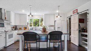 A modern kitchen with white cabinets, an island with two black chairs, pendant lights, stainless steel appliances, a window above the sink, and a sliding barn door leading to a pantry.
