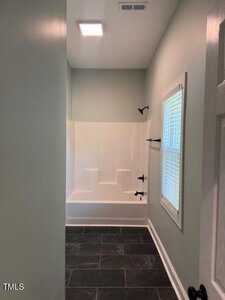 A bathroom with light green walls, a white bathtub and shower combo with black fixtures, a window with blinds, and dark tile flooring. A ceiling light and air vent are visible above.