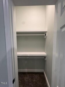 A small walk-in closet with light-colored walls, two white shelves mounted on the back wall, and dark carpeted flooring. The closet door is partially open.