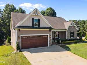 A single-story suburban house with beige siding, stone accents, a prominent gable, and a two-car garage with wooden-style doors. The front yard is landscaped with grass and small shrubs. An American flag hangs near the entryway.