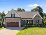 Located at 3658 Genesis Lane in Wake Forest, this charming one-story home features a gable roof, stone and siding exterior, two-car garage, inviting front porch with chairs, and a well-kept lawn. An American flag hangs near the porch amid shady trees.