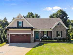 Located at 3658 Genesis Lane in Wake Forest, this charming one-story home features a gable roof, stone and siding exterior, two-car garage, inviting front porch with chairs, and a well-kept lawn. An American flag hangs near the porch amid shady trees.